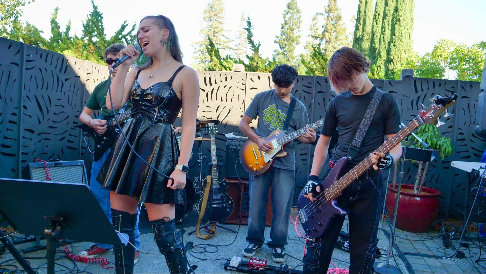 A four-piece rock band performing outdoors against a decorative black fence. A female lead singer in a black faux-leather dress and tall boots sings into a microphone, flanked by two guitarists and a bassist in punk-inspired attire.