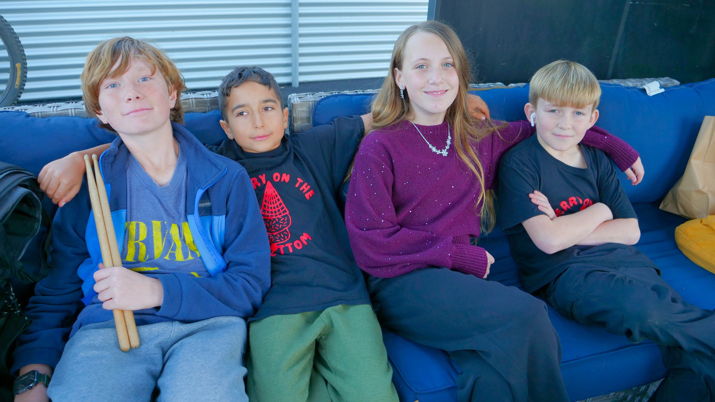 Four children sitting together on a blue outdoor couch with their arms around each other, smiling at the camera. One child on the left holds drumsticks and wears a blue jacket and graphic t-shirt, while two children wear black shirts with red lettering. The child in the middle wears a purple sweater and necklace. A bicycle wheel and bags are visible in the background, suggesting an outdoor or patio setting.