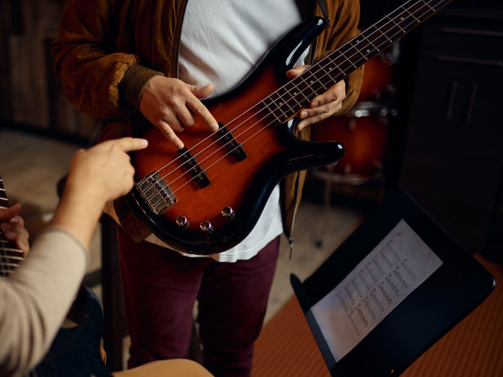 Beginner bass guitar lesson showing hands demonstrating finger placement and technique on electric bass during instruction, focusing on proper playing form and guidance.