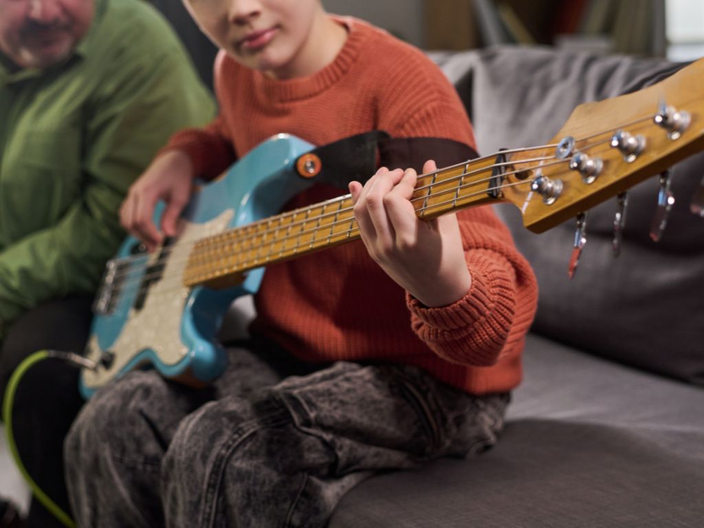 Beginner bass guitar lesson showing a child learning bass guitar at home, focusing on hand positioning and finger technique with instructor guidance in a relaxed practice setting.